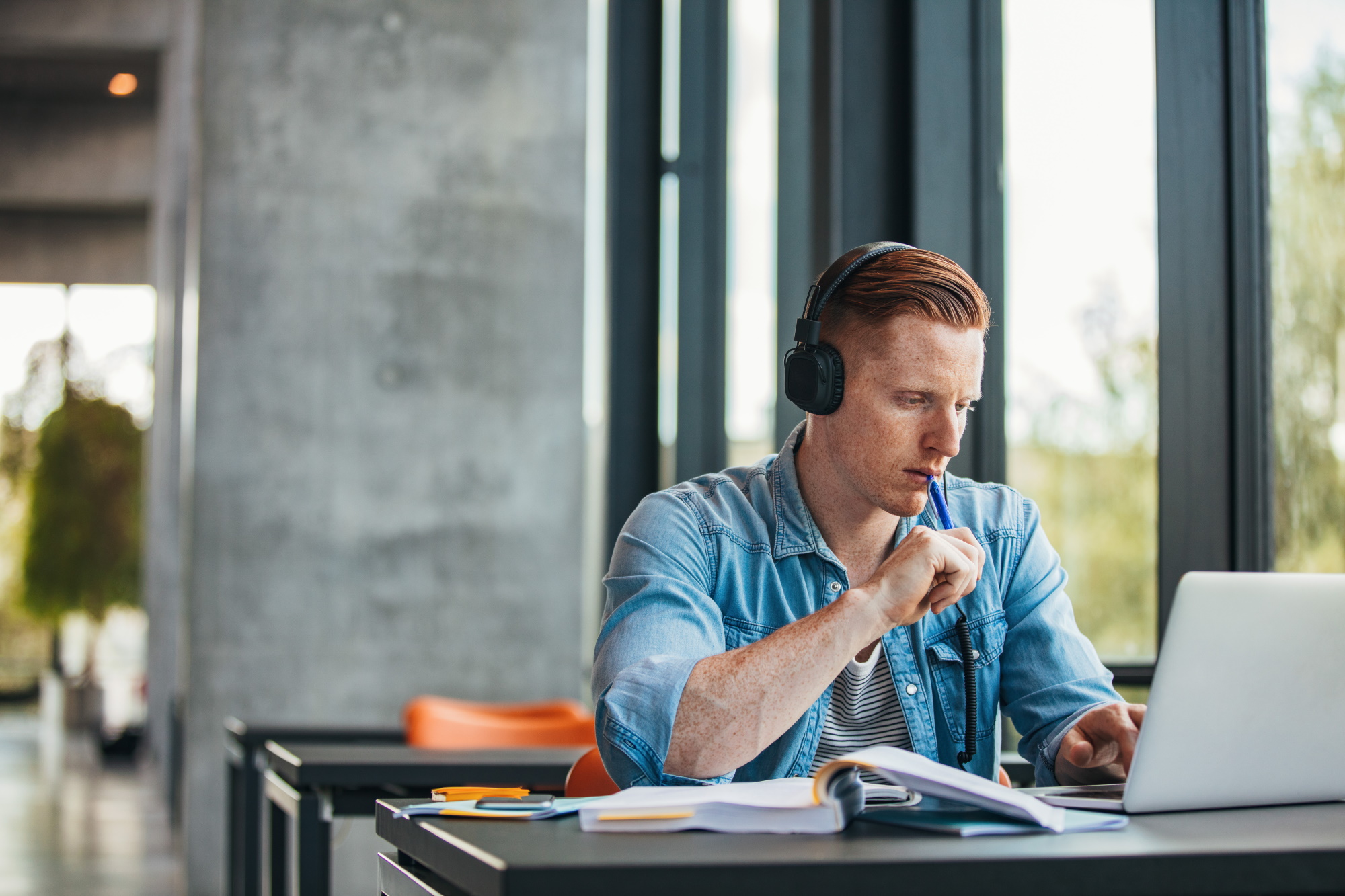 Student mit Kopfhörern sitzt an einem Tisch und arbeitet fokussiert am Laptop, mit Büchern und Notizen vor sich.