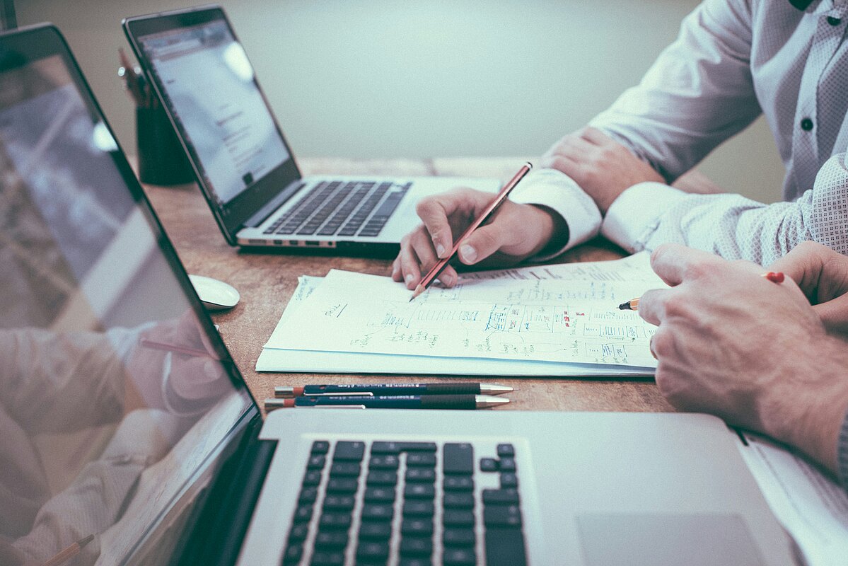  Zwei Personen arbeiten an Laptops, überprüfen Papiere und machen Notizen. Fokus auf die Hände, die einen Bleistift und einen Stift halten.
