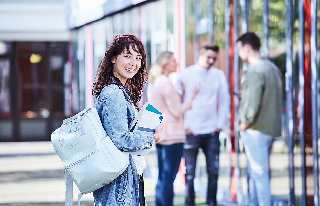 Junge Studentin mit lockigem Haar, Jeansjacke und Rucksack hält Bücher in der Hand und lächelt, während im Hintergrund Studierende miteinander sprechen.