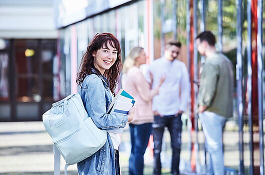 Junge Studentin mit lockigem Haar, Jeansjacke und Rucksack hält Bücher in der Hand und lächelt, während im Hintergrund Studierende miteinander sprechen.