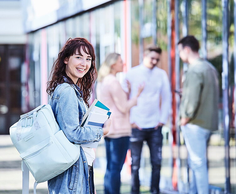 Public Management studieren Junge Studentin mit lockigem Haar, Jeansjacke und Rucksack hält Bücher in der Hand und lächelt, während im Hintergrund Studierende miteinander sprechen.