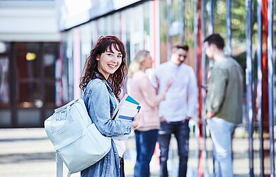 Junge Studentin mit lockigem Haar, Jeansjacke und Rucksack hält Bücher in der Hand und lächelt, während im Hintergrund Studierende miteinander sprechen.