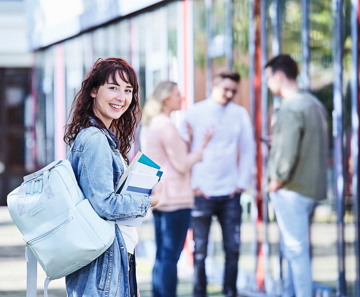 Junge Studentin mit lockigem Haar, Jeansjacke und Rucksack hält Bücher in der Hand und lächelt, während im Hintergrund Studierende miteinander sprechen.