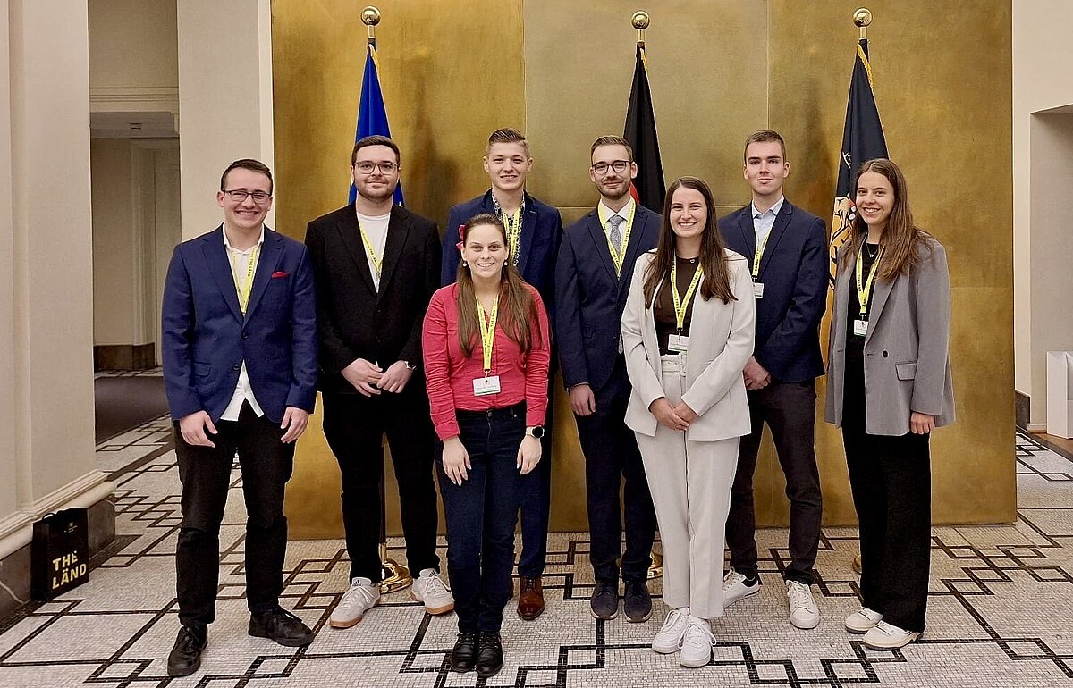 Gruppenfoto der besten Bachelorabsolvent:innen (drei Frauen, fünf Männer) beim Empfang im Staatsministerium, stehend vor einer goldenen Wand und Fahnen.