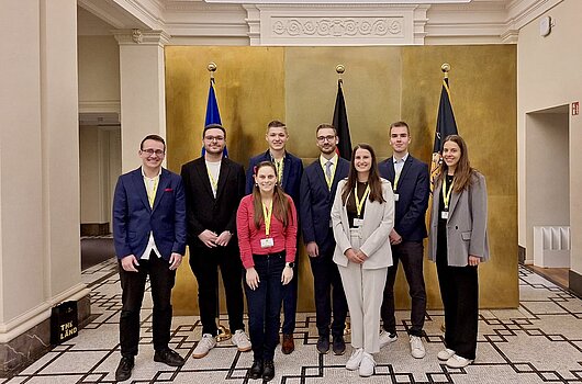 Gruppenfoto der besten Bachelorabsolvent:innen (drei Frauen, fünf Männer) beim Empfang im Staatsministerium, stehend vor einer goldenen Wand und Fahnen.