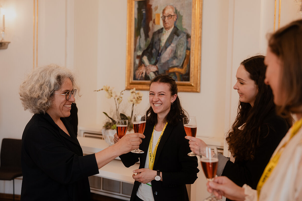 Empfang im Staatsministerium Ministerialdirigentin Annegret Breitenbücher stößt in einem eleganten Raum mit Sektgläsern mit drei Absolventinnen an. Sie lächeln. Im Hintergrund ein Porträt und eine Orchidee.