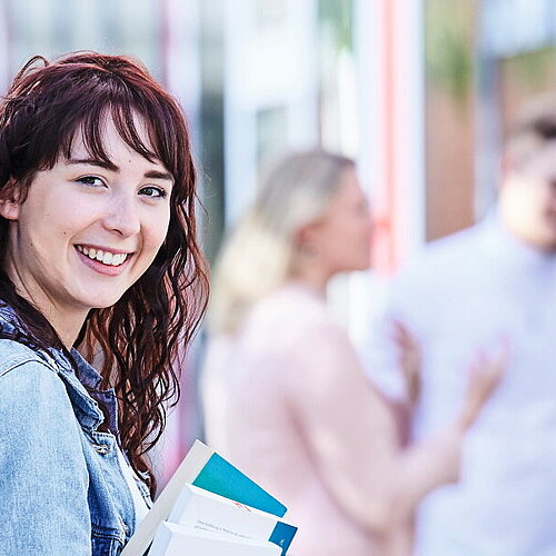 Junge Studentin mit lockigem Haar, Jeansjacke und Rucksack hält Bücher in der Hand und lächelt, während im Hintergrund Studierende miteinander sprechen.