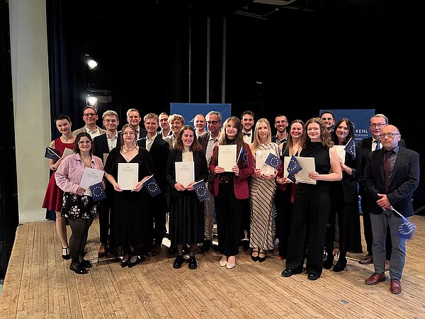 Stolze Absolventen des Master Europäischen Verwaltungsmanagement (MEPA) posieren mit Urkunden und EU-Flaggen.