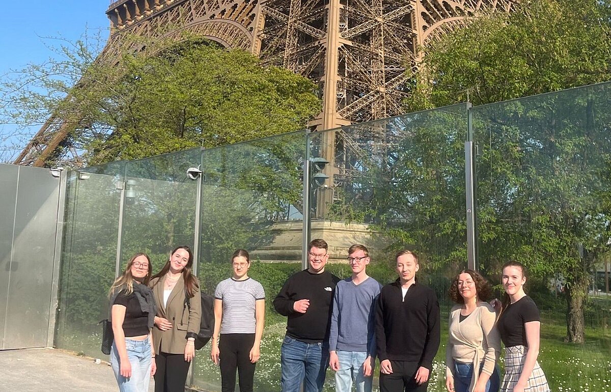 Sieben Studierende stehen vor dem Eiffelturm an einem sonnigen Tag mit blauem Himmel und grünen Bäumen im Hintergrund.