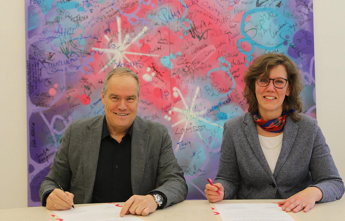 Eckart Würzner und Dr. Iris Eliisa Rauskala unterzeichnen eine Absichtserklärung vor einem bunten Kunstwerk in der Stadt Heidelberg.