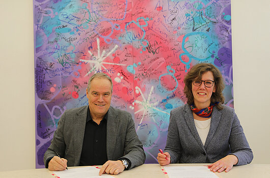 Eckart Würzner und Dr. Iris Eliisa Rauskala unterzeichnen eine Absichtserklärung vor einem bunten Kunstwerk in der Stadt Heidelberg.