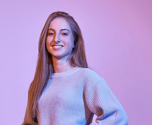 Rentenversicherung studieren Porträt einer jungen Studentin der Rentenversicherung mit langen Haaren und Strickpullover vor einem lila Hintergrund, lächelnd und mit selbstbewusster Haltung.