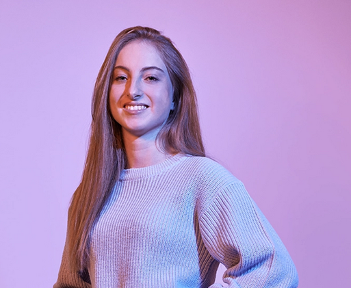 Porträt einer jungen Studentin der Rentenversicherung mit langen Haaren und Strickpullover vor einem lila Hintergrund, lächelnd und mit selbstbewusster Haltung.