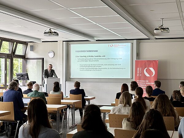 Antrittsvorlesung Prof. Dr. Philipp Kauffmann Professor Kauffmann hält eine Vorlesung zu „Der Tatbestand der Steuerhinterziehung in Wissenschaft und Praxis“ vor einem Publikum, mit Präsentation und HVF-Banner.