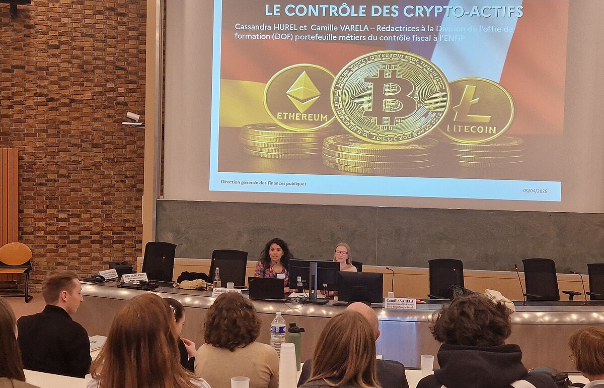 Studierende sitzen vor einer Leinwand mit der Aufschrift "LE CONTRÔLE DES CRYPTO-ACTIFS" und Bildern von Ethereum-, Bitcoin- und Litecoin-Logos. Zwei Frauen präsentieren vorne.