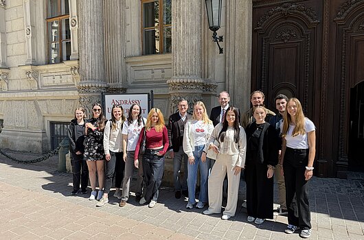 Exkursionsgruppe vor der Andrássy Universität Budapest Eine Gruppe von zwölf Studierenden und zwei Professoren steht in zwei Reihen vor einem verzierten, hellen Steingebäude mit einem Schild, das "ANDRÁSSY U" zeigt, und einer großen Holztür.