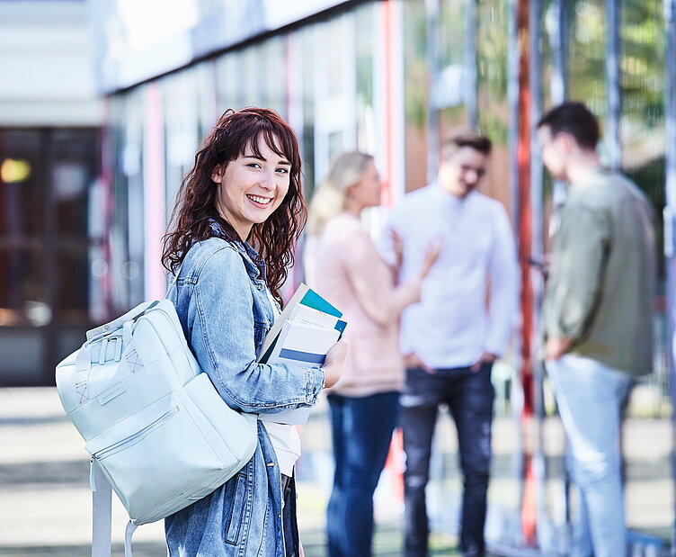 Junge Studentin mit lockigem Haar, Jeansjacke und Rucksack hält Bücher in der Hand und lächelt, während im Hintergrund Studierende miteinander sprechen.