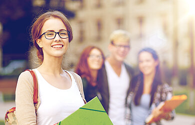 Junge Studentin mit Brille und Ordnern lächelt in die Kamera, im Hintergrund weitere Studierende auf einem sonnigen Campus.