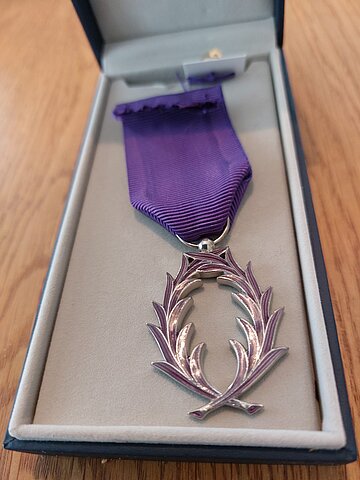 Nahaufnahme eines Ordens der „Palmes Académiques“ in einer offenen Schatulle. Der Orden besteht aus einem silberfarbenen Lorbeerkranz, der an einem violettfarbenen Band hängt. Die Schatulle liegt auf einem Holzuntergrund.