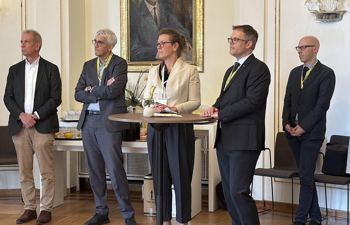 Dr. Rauskala (Mitte) und weitere Gäste verfolgen aufmerksam die Ordensverleihung an Prof. Dr. Angelika Dölker. Die Szene spielt in einem feierlich gestalteten Raum mit klassischem Ambiente.