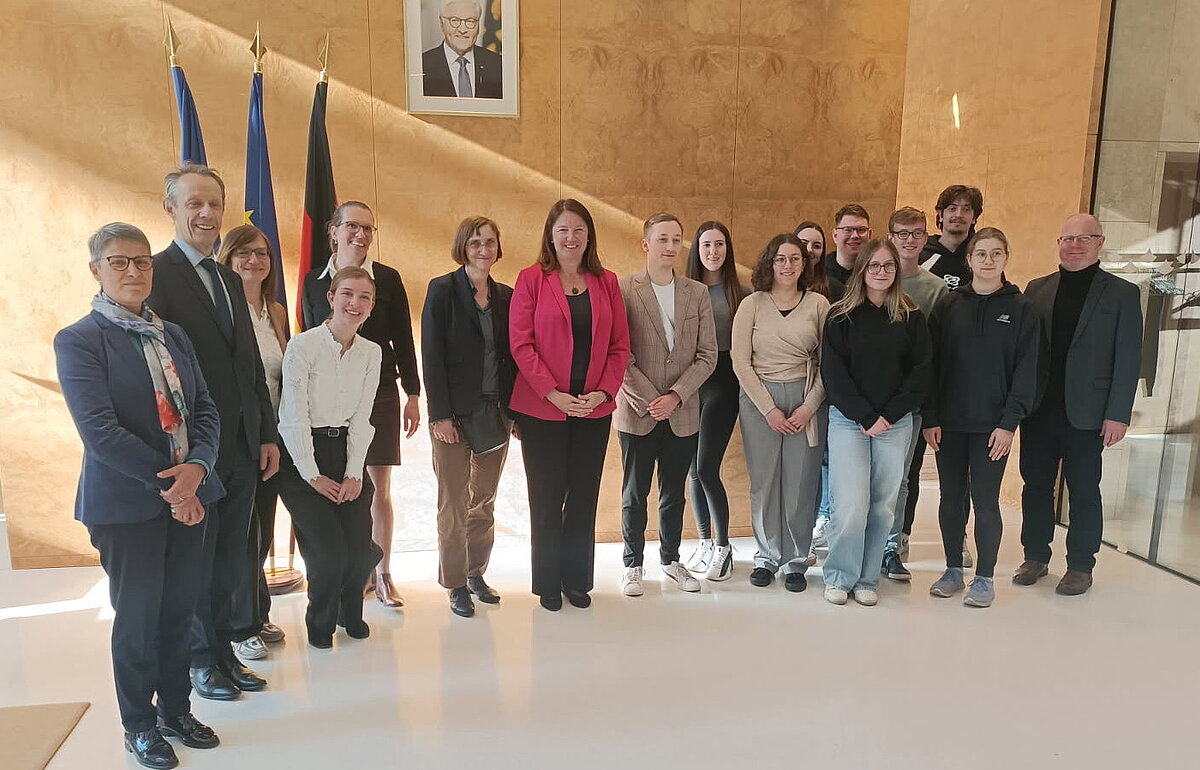 Eine Gruppe von Studierenden und Erwachsenen posiert für ein Gruppenfoto im Finanzministerium mit Flaggen und einem Porträt an der Wand.