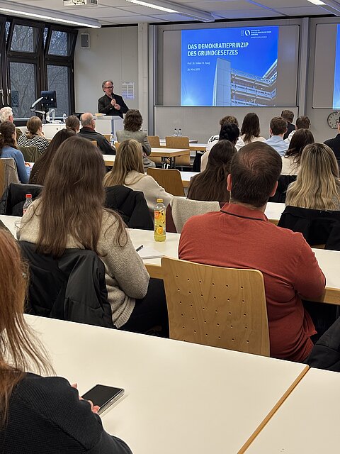 Ein Mann - Prof. Dr. Volker M. Haug - steht hinter einem Rednerpult in einem gefüllten Vorlesungsraum. Hinter ihm ist eine Leinwand, sie zeigt eine Präsentation mit dem Titel "Das Demokratieprinzip des Grundgesetzes"