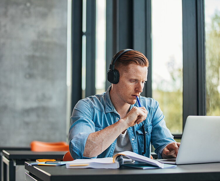 Student mit Kopfhörern sitzt an einem Tisch und arbeitet fokussiert am Laptop, mit Büchern und Notizen vor sich.