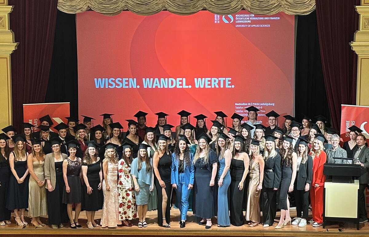 Der Abschlussjahrgang 2025 des Studiengangs Rentenversicherung der Hochschule Ludwigsburg posiert in Absolventenhüten auf der Bühne.