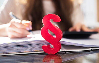Rotes Paragraphenzeichen vor einer studierenden Person mit aufgeschlagenen Unterlagen, Stift und Taschenrechner auf dem Tisch.