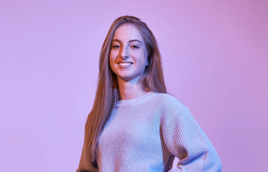Porträt einer jungen Studentin der Rentenversicherung mit langen Haaren und Strickpullover vor einem lila Hintergrund, lächelnd und mit selbstbewusster Haltung.