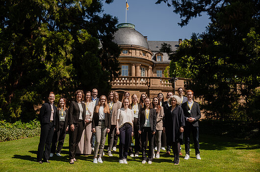 Gruppenfoto der besten Absolventen vor der Villa Reitzenstein Eine Gruppe junger Frauen und Männer steht auf einem grünen Rasen vor einem großen, hellbraunen Gebäude mit einer grauen Kuppel und einer Baden-Württemberg-Flagge auf dem Dach. Es handelt sich um die Villa Reitzenstein. Sie lächeln in die Kamera. Bäume rahmen die Szene.