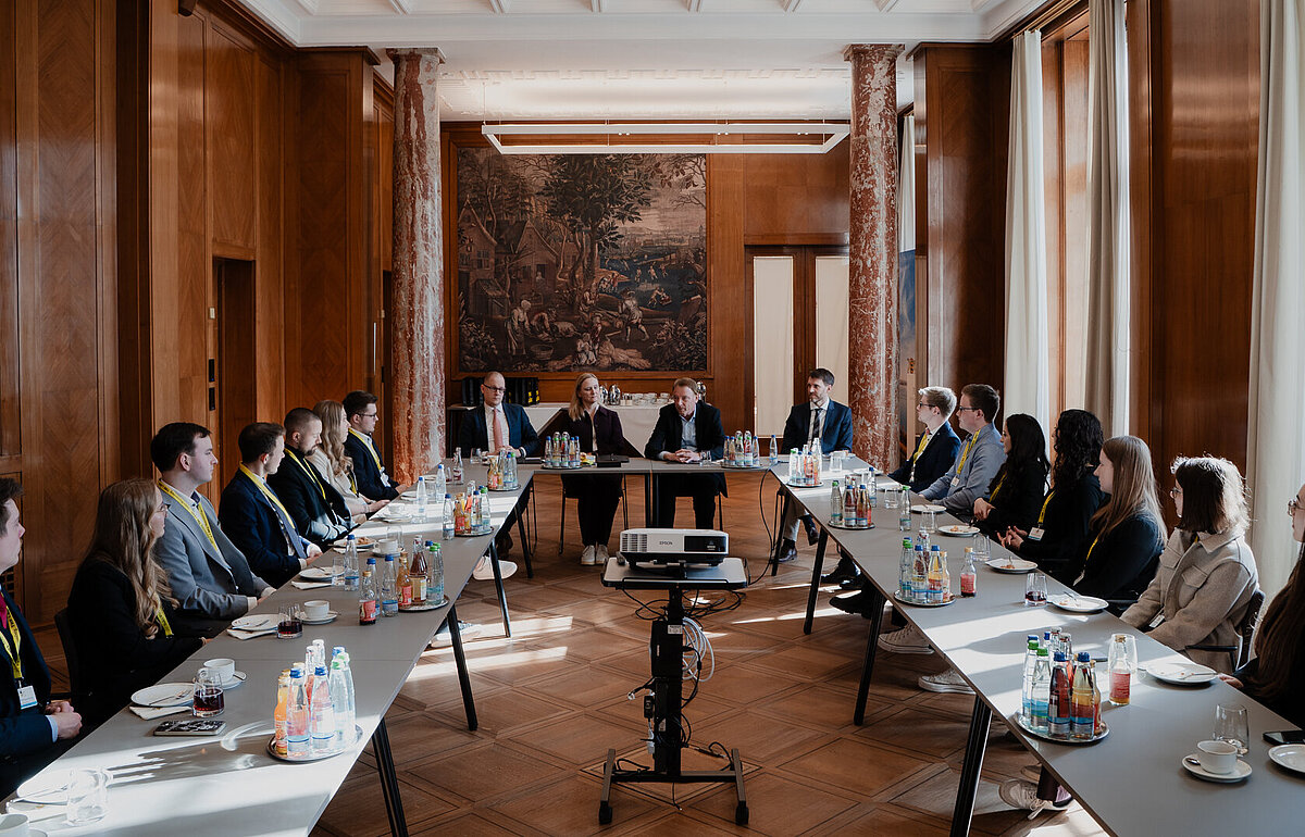 Gruppenbild am Tisch sitzender Absolventinnen und Absolventen, die dem Vortrag zuhören