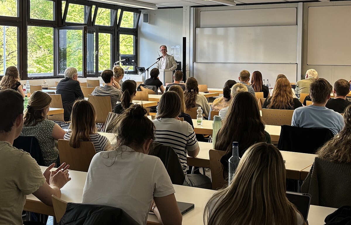 Volker Haug steht an einem Rednerpult und liest aus dem Buch "Kompass politische Kultur" von Ulrich Müller vor einem aufmerksamen Publikum.