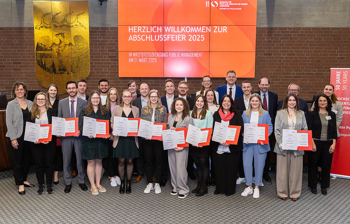 Eine Gruppe von etwa 25 Personen posiert für ein Abschlussfoto bei der Abschlussfeier 2025 des Masterstudiengangs Public Management. Die Absolventinnen und Absolventen stehen in der vorderen Reihe und halten ihre Urkunden mit roten Mappen in den Händen. Hinter ihnen stehen weitere Personen, vermutlich Dozierende und Hochschulvertreter. Im Hintergrund befindet sich eine große digitale Anzeigetafel mit der Aufschrift „Herzlich willkommen zur Abschlussfeier 2025“ sowie das Logo der Hochschule für öffentliche Verwaltung und Finanzen Ludwigsburg.