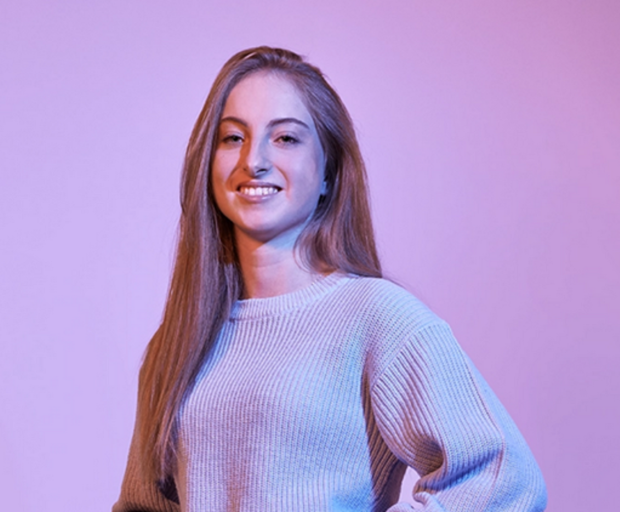 Porträt einer jungen Studentin der Rentenversicherung mit langen Haaren und Strickpullover vor einem lila Hintergrund, lächelnd und mit selbstbewusster Haltung.