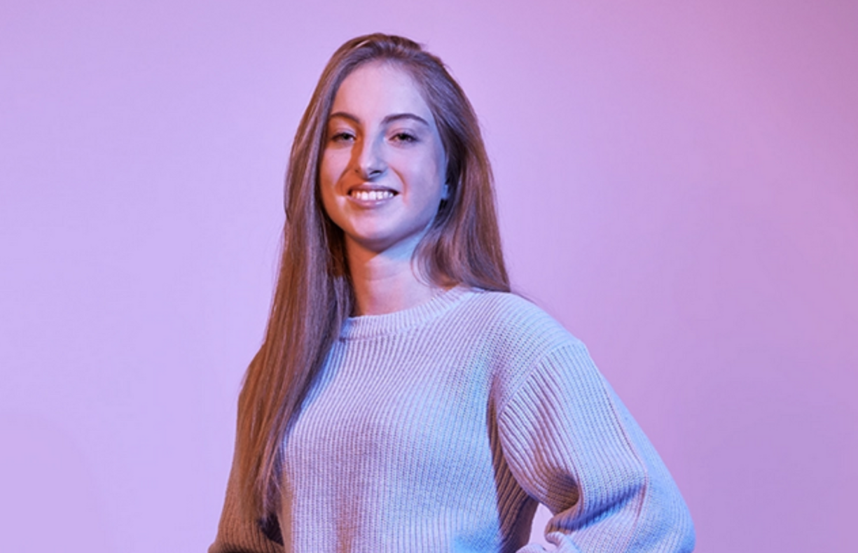 Porträt einer jungen Studentin der Rentenversicherung mit langen Haaren und Strickpullover vor einem lila Hintergrund, lächelnd und mit selbstbewusster Haltung.