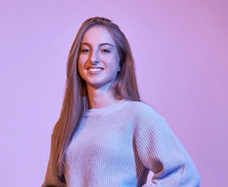 Rentenversicherung studieren Porträt einer jungen Studentin der Rentenversicherung mit langen Haaren und Strickpullover vor einem lila Hintergrund, lächelnd und mit selbstbewusster Haltung.