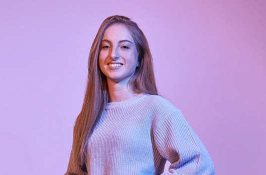 Porträt einer jungen Studentin der Rentenversicherung mit langen Haaren und Strickpullover vor einem lila Hintergrund, lächelnd und mit selbstbewusster Haltung.