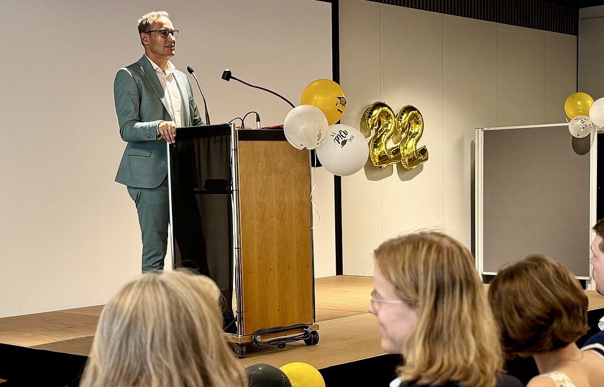 William Schmitt spricht bei der Abschlussfeier an einem Pult mit Ballons im Hintergrund, während ein Publikum von hinten zu sehen ist.