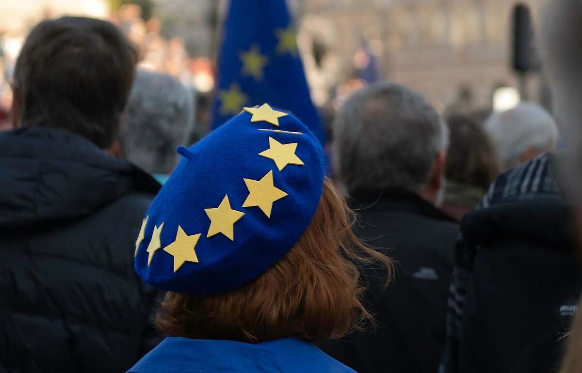 Frau mit blauem EU-Barett in einer Menschenmenge bei einer pro-europäischen Demonstration auf dem Trafalgar Square in London, im Hintergrund sind Gebäude und eine EU-Flagge sichtbar.