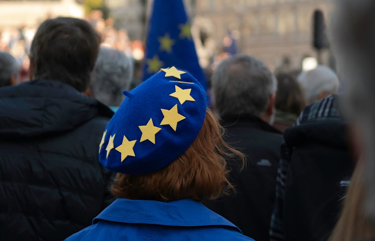 Frau mit blauem EU-Barett in einer Menschenmenge bei einer pro-europäischen Demonstration auf dem Trafalgar Square in London, im Hintergrund sind Gebäude und eine EU-Flagge sichtbar.