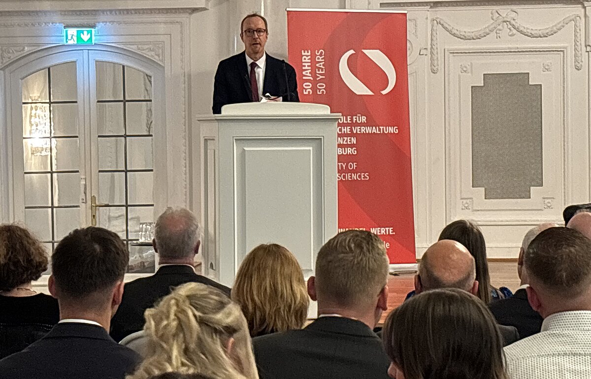 Christian Järkel vom Finanzministerium hält eine Rede von einem Rednerpult vor einem Publikum bei der Bachelorfeier.