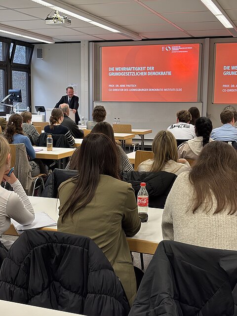 250326_Demokratie_Pautsch Ein Mann - Prof. Dr. Arne Pautsch - steht hinter einem Rednerpult in einem Seminarraum. Hinter ihm sind zwei Leinwände zu sehen, auf diesen steht auf rotem Hintergrund "Die Wehrhaftigkeit der Demokratie"