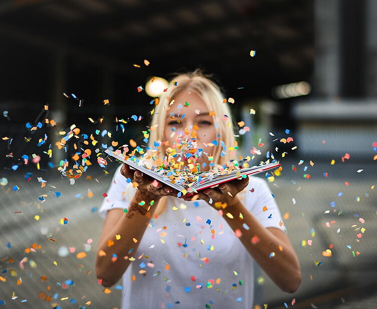 Frau pustet buntes Konfetti von einem aufgeschlagenen Buch, Symbol für Lebensfreude beim Kongress für Positive Psychologie.