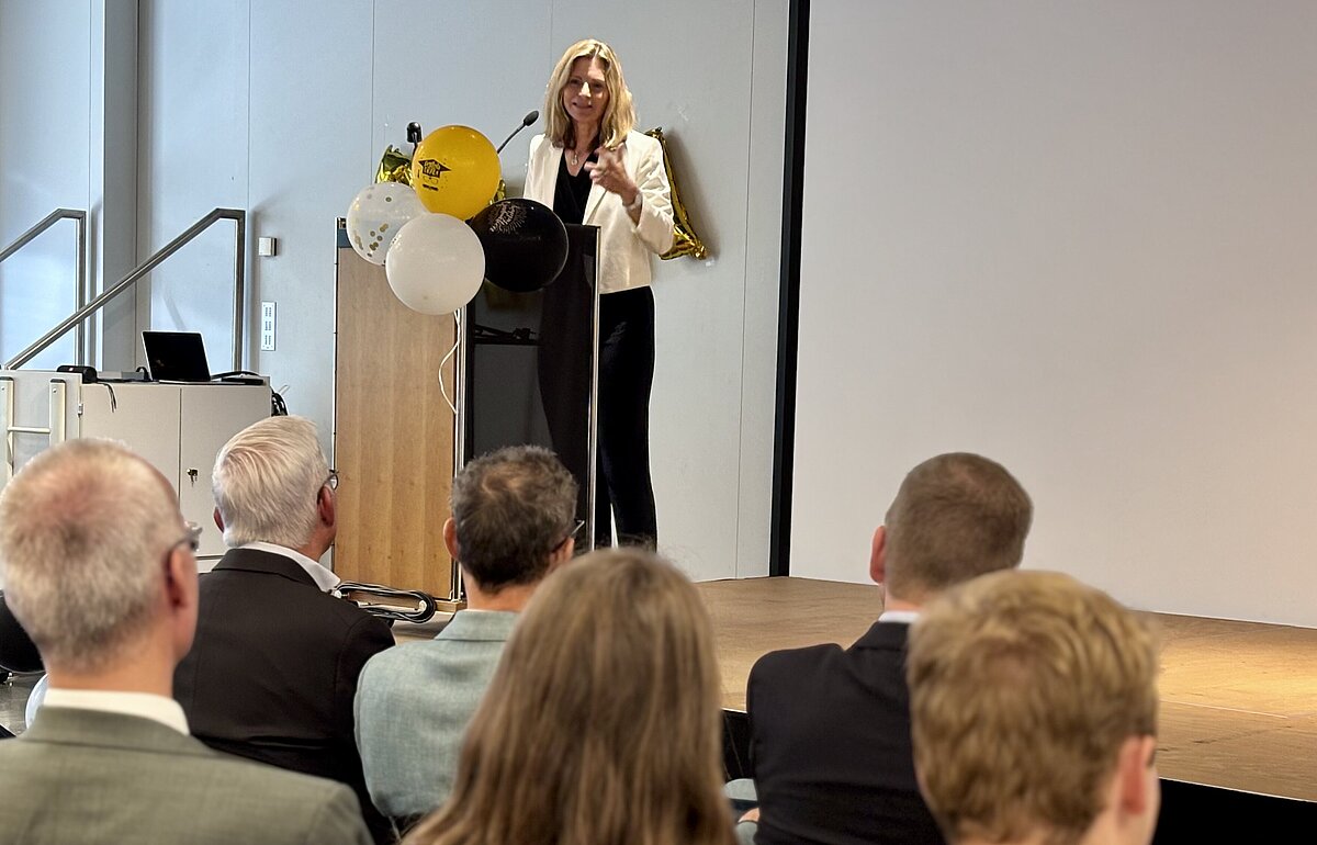 Heidi Schmid spricht von einem Rednerpult aus vor einem Publikum. Dekoration mit Ballons und Buchstaben im Hintergrund.