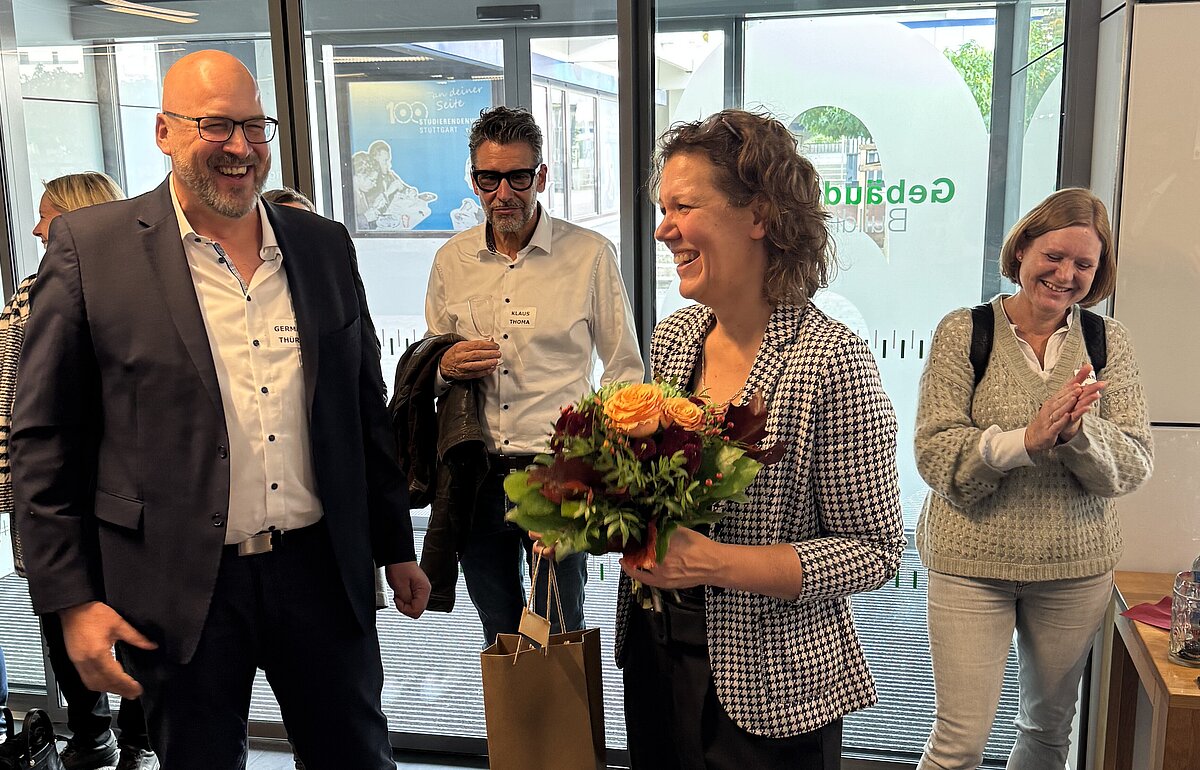  German Thüry (Organisator) und Rektorin Dr. Iris Rauskala (mit Blumenstrauß) lächeln im Foyer der HVF Ludwigsburg, umgeben von Teilnehmern.