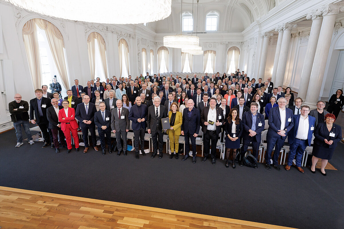 Gruppenfoto bei der Unterzeichnung von HoFV III Gruppenfoto in einem prunkvollen Saal mit hohen Decken, grossen Fenstern und eleganten Kronleuchtern. Im Vordergrund stehen Mitglieder der Landesregierung sowie Rektorinnen, Rektoren, Präsidentinnen, Präsidenten, Kanzlerinnen und Kanzler der baden-württembergischen Hochschulen zusammen mit weiteren Gästen. Einige Personen halten Dokumente in den Händen, was auf die feierliche Unterzeichnung der HoFV III hinweist.