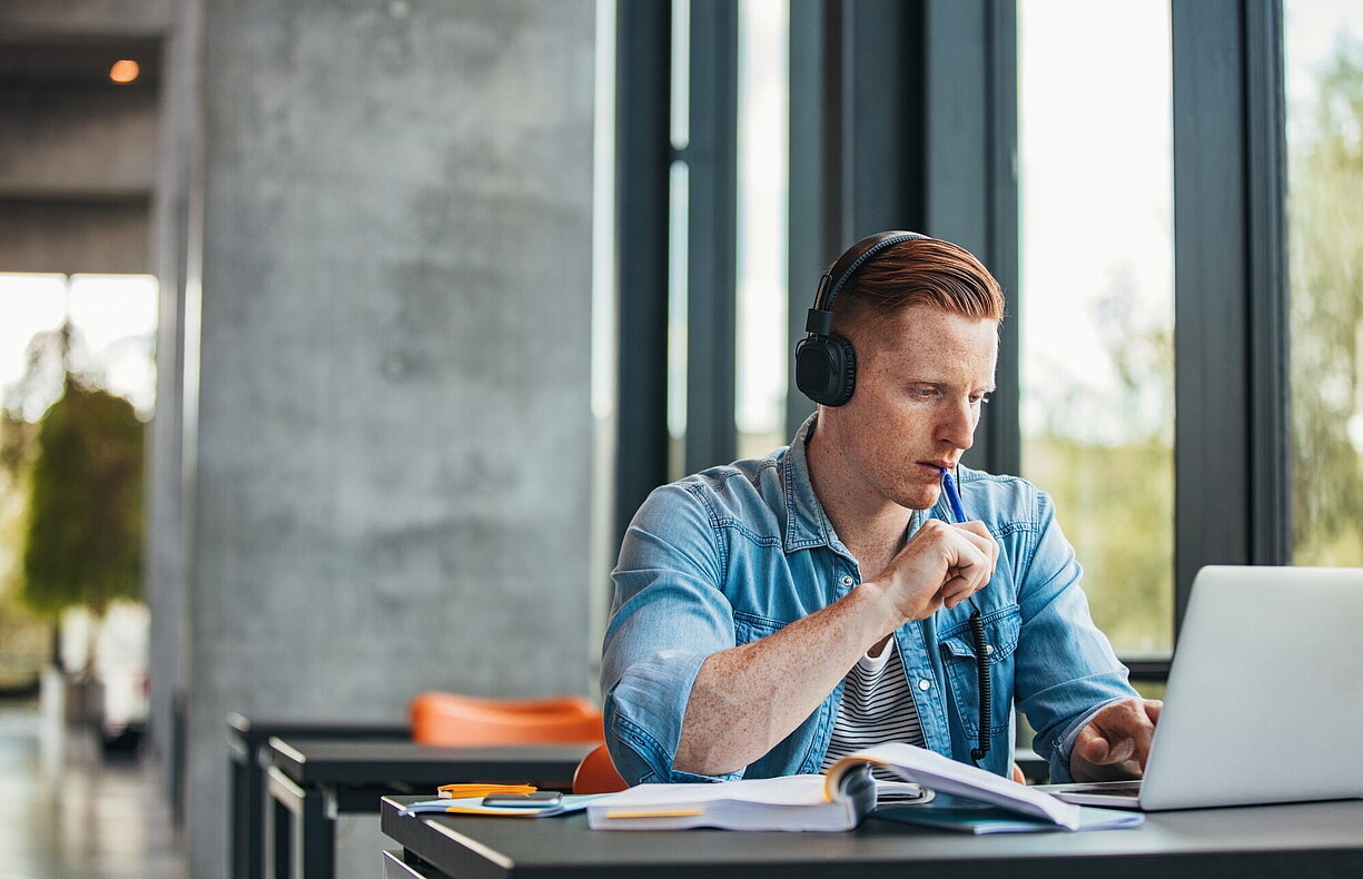 Student mit Kopfhörern sitzt an einem Tisch und arbeitet fokussiert am Laptop, mit Büchern und Notizen vor sich.