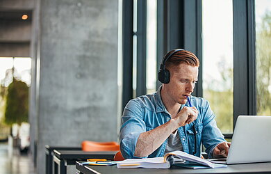 Student mit Kopfhörern sitzt an einem Tisch und arbeitet fokussiert am Laptop, mit Büchern und Notizen vor sich.