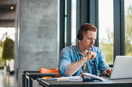 Student mit Kopfhörern sitzt an einem Tisch und arbeitet fokussiert am Laptop, mit Büchern und Notizen vor sich.